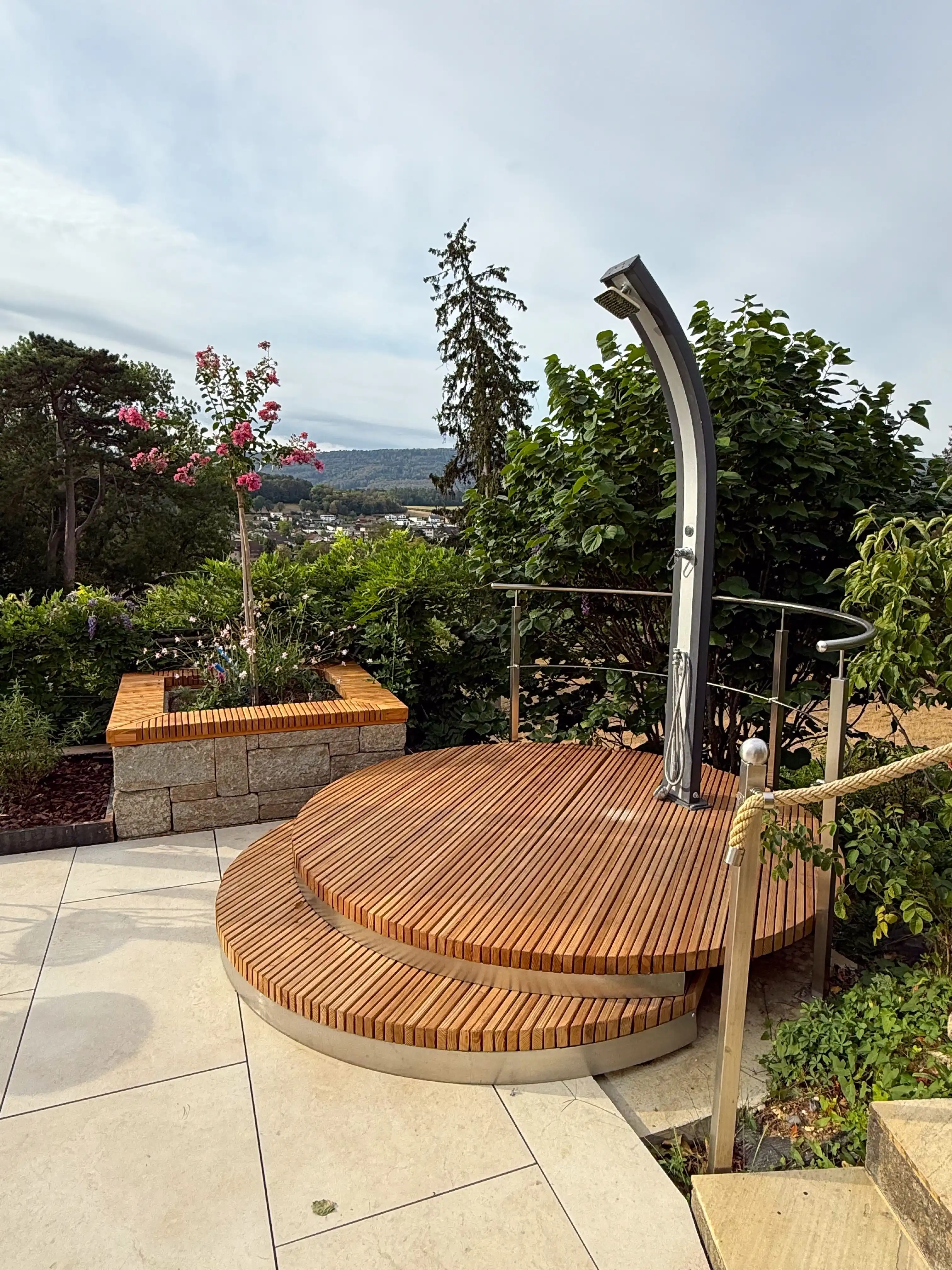Second bac en pierre coiffé de bois, planté d'un arbuste en fleurs, à côté d'une terrasse ronde en bois